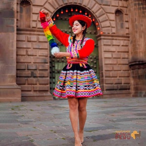 Op de afbeelding: Een vrouw in traditionele Peruaanse kleding, waaronder een rode hoed en een felgekleurde jurk met patronen. De outfit heeft kleurrijk borduurwerk en een regenboogkleurige kwast. De foto is buiten gemaakt.