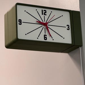 Pode incluir: Um relógio de parede retangular com mostrador branco, marcadores de hora pretos e ponteiros vermelhos. O relógio tem uma moldura verde escura e está montado na parede. O mostrador do relógio apresenta os números 3, 6, 9 e 12.