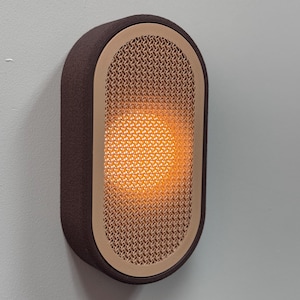 Peut inclure: Applique murale moderne avec un cadre rectangulaire marron. Le centre pr&eacute;sente un ovale beige textur&eacute;, &eacute;mettant une lumi&egrave;re orange chaude. L'applique est fix&eacute;e sur un mur gris clair, cr&eacute;ant une ambiance minimaliste.