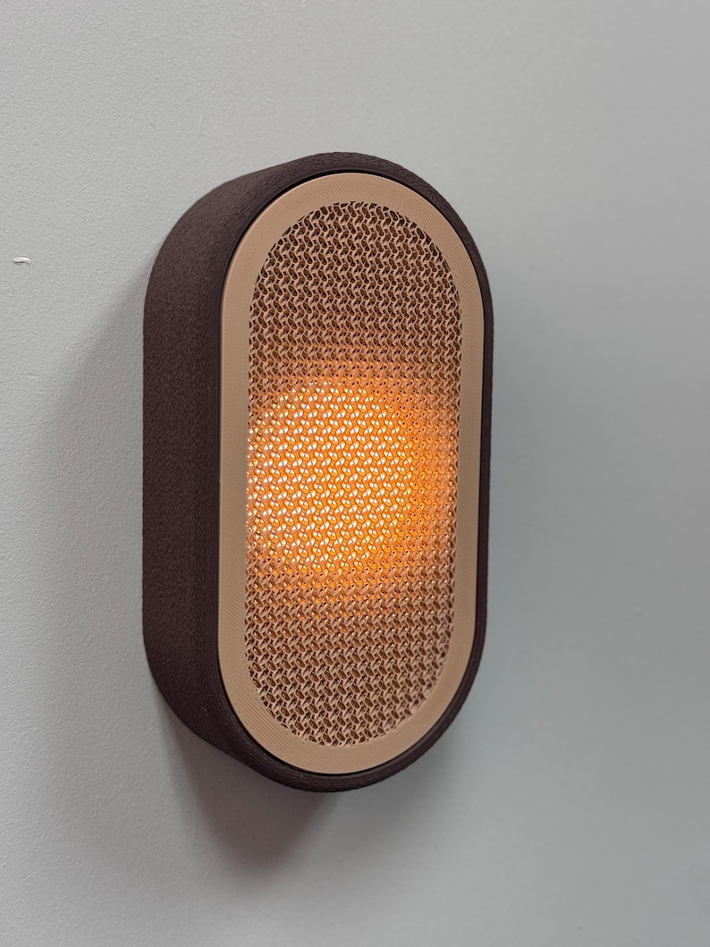 Peut inclure: Applique murale moderne avec un cadre rectangulaire marron. Le centre pr&eacute;sente un ovale beige textur&eacute;, &eacute;mettant une lumi&egrave;re orange chaude. L'applique est fix&eacute;e sur un mur gris clair, cr&eacute;ant une ambiance minimaliste.