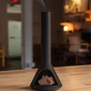 Op de afbeelding: Een zwarte tafelhaard met een rookpluim die uit de schoorsteen opstijgt. De haard heeft een realistisch houtontwerp en is een decoratief stuk.