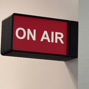 May include: A rectangular, black-framed sign with a red background. The words "ON AIR" are displayed in large, white capital letters. The sign is mounted on a light-colored wall, indicating its use in a broadcast or recording environment.