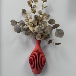 Puede incluir: Un jarrón acanalado de color coral con un cuello delgado, que contiene ramas de eucalipto artificiales con hojas redondas, verdes y amarillas apagadas. El jarrón está montado en una pared blanca, creando una exhibición decorativa simple y moderna.