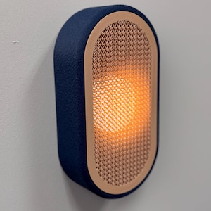 Peut inclure: Applique murale bleu marine avec une lueur orange chaude. L'applique a une forme rectangulaire arrondie avec une fa&ccedil;ade textur&eacute;e en cuivre tiss&eacute; et un ext&eacute;rieur bleu marine semblable &agrave; du tissu. Mont&eacute;e sur un mur gris clair.
