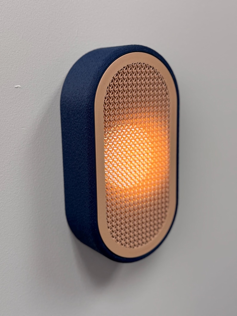 Peut inclure: Applique murale bleu marine avec une lueur orange chaude. L'applique a une forme rectangulaire arrondie avec une fa&ccedil;ade textur&eacute;e en cuivre tiss&eacute; et un ext&eacute;rieur bleu marine semblable &agrave; du tissu. Mont&eacute;e sur un mur gris clair.