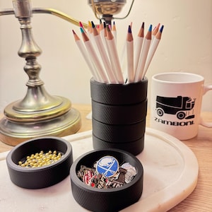 May include: A desk scene featuring a black pencil holder filled with white pencils with colored tips. Two small black trays hold gold beads and pins. A white mug with a black graphic and the word "Zamboni" is also present.