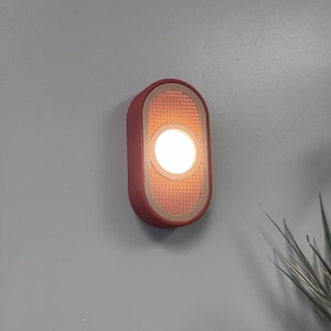 May include: A contemporary wall light with a red-orange casing and a textured, copper-toned centre. The light source is a bright, circular bulb. The fixture is mounted on a grey wall.