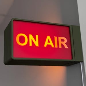 Puede incluir: Un letrero rectangular "ON AIR" con fondo rojo y letras amarillas iluminadas. El letrero está encerrado en un marco verde oscuro y montado en una pared. El letrero se utiliza en un estudio de grabación o entorno de transmisión.