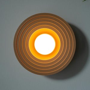 May include: A circular wooden wall light with a series of concentric rings and a warm, bright glow. The light fitting is mounted on a grey wall, creating a modern, minimalist look. The light is approximately 30 cm in diameter.