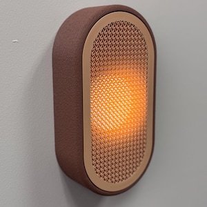 Peut inclure: Applique murale ovale marron avec un ext&eacute;rieur textur&eacute; et un int&eacute;rieur en forme de treillis. L'int&eacute;rieur brille d'une lumi&egrave;re orange chaude. L'applique est fix&eacute;e sur un mur gris clair.