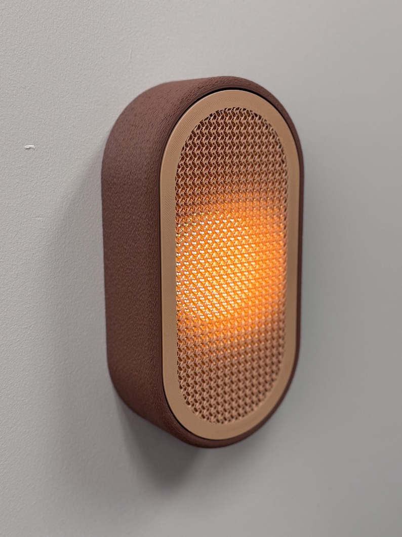 Peut inclure: Applique murale ovale marron avec un ext&eacute;rieur textur&eacute; et un int&eacute;rieur en forme de treillis. L'int&eacute;rieur brille d'une lumi&egrave;re orange chaude. L'applique est fix&eacute;e sur un mur gris clair.