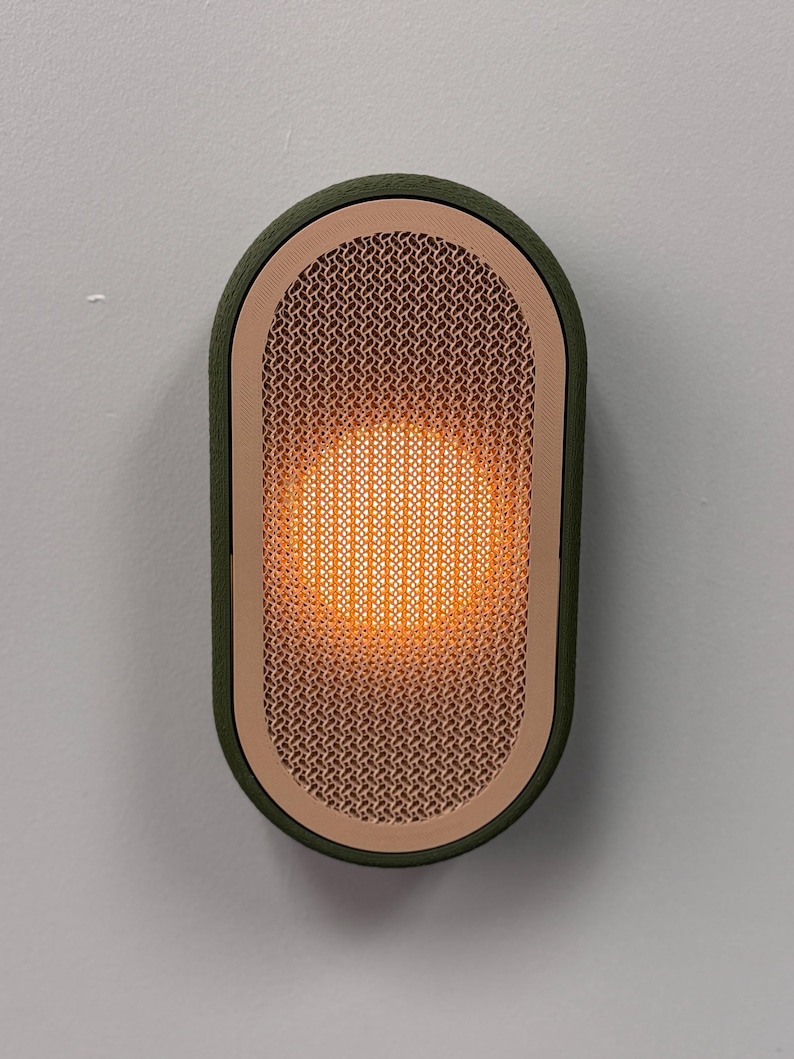 Peut inclure: Applique murale ovale avec un cadre vert et une fa&ccedil;ade en treillis de couleur cuivre. Le centre brille d'une lumi&egrave;re orange chaude. L'applique est fix&eacute;e sur un mur gris clair.