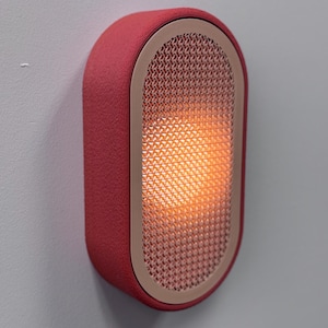 Peut inclure: Applique murale ovale rouge avec une surface textur&eacute;e. Le luminaire pr&eacute;sente un panneau avant en forme de grille, d&eacute;gageant une lueur orange chaude. Un cadre beige clair entoure la grille. Fix&eacute; sur un mur gris clair. Mesure environ 20 cm de haut.