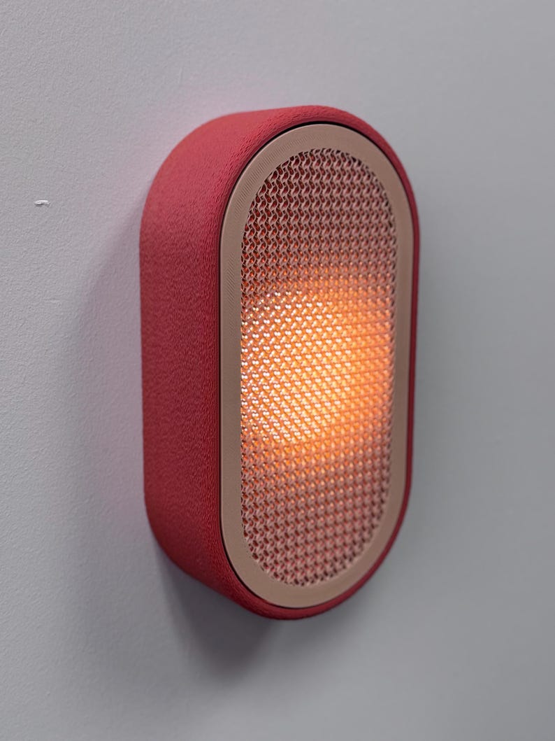 Peut inclure: Applique murale ovale rouge avec une surface textur&eacute;e. Le luminaire pr&eacute;sente un panneau avant en forme de grille, d&eacute;gageant une lueur orange chaude. Un cadre beige clair entoure la grille. Fix&eacute; sur un mur gris clair. Mesure environ 20 cm de haut.