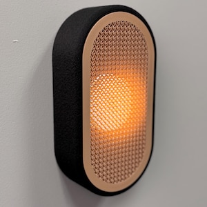 Peut inclure: Applique murale ovale avec un cadre noir textur&eacute; et une fa&ccedil;ade en maille de couleur cuivre. La lumi&egrave;re &agrave; l'int&eacute;rieur de l'applique &eacute;met une lueur orange chaude. L'applique est fix&eacute;e sur un mur gris clair.