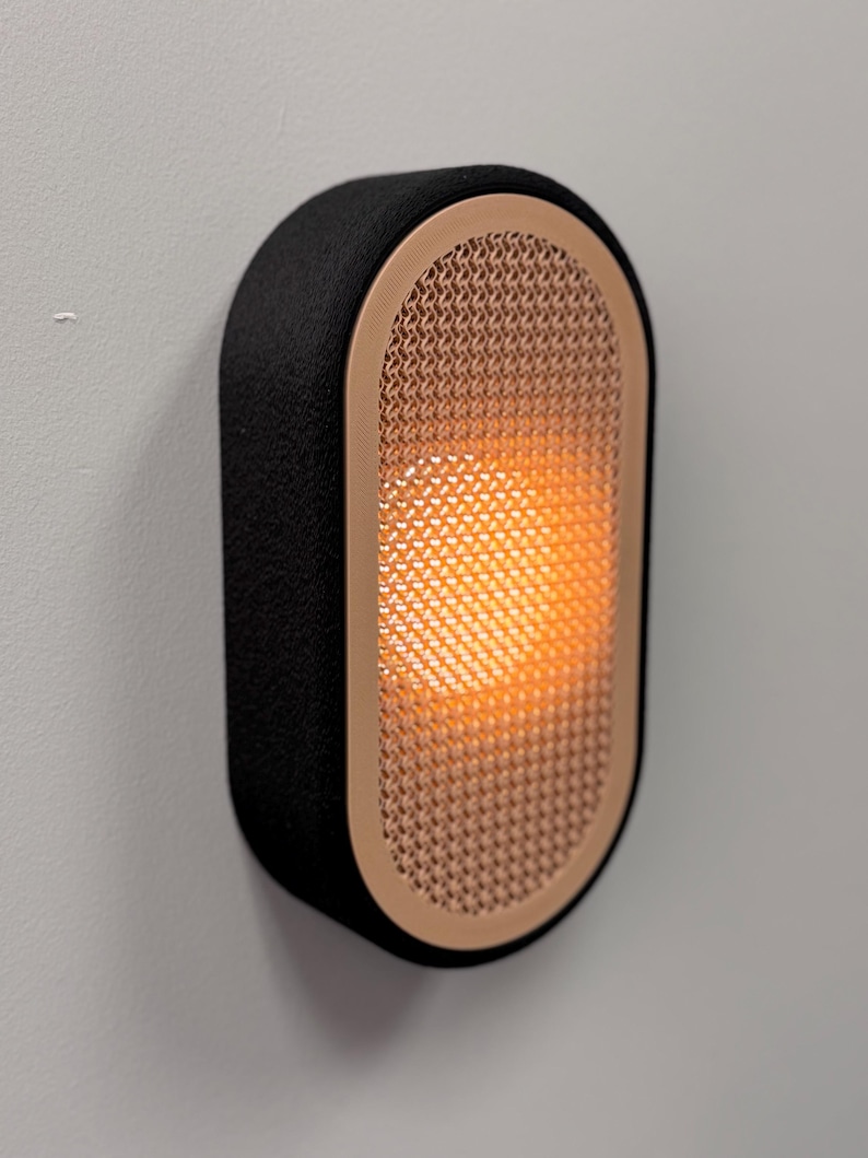 Peut inclure: Applique murale ovale avec un cadre noir textur&eacute; et une fa&ccedil;ade en maille de couleur cuivre. La lumi&egrave;re &agrave; l'int&eacute;rieur de l'applique &eacute;met une lueur orange chaude. L'applique est fix&eacute;e sur un mur gris clair.