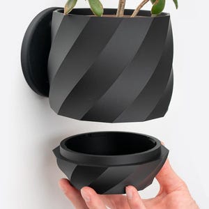 Könnte beinhalten: Ein schwarzer, geometrischer Wandtopf mit abnehmbarem Boden. Der Topf besteht aus glattem, mattem Kunststoff und hat ein einzigartiges, gedrehtes Design.