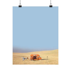 May include: A vintage orange camper van parked in a field of tall grass with a person standing in the distance. The sky is a light blue color.