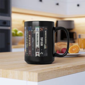 May include: A black ceramic coffee mug featuring a design of stacked VHS tapes. The tapes include titles like "Platoon," "Red Dawn," "Saving Private Ryan," "Top Gun," and "Windtalkers." The mug has a large handle and is set on a wooden surface.