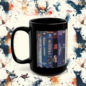 May include: Black ceramic mug featuring a collage of VHS tapes, including "The Addams Family," "Back to the Future," and "Beetlejuice." The mug has a large handle and a glossy finish. The background has a pattern of watercolor animals.
