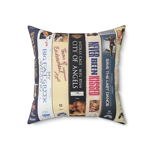 May include: Square throw pillow featuring a collage of VHS movie covers. Titles include "My Big Fat Greek Wedding," "City of Angels," and "Never Been Kissed." The pillow has a white background and a soft, textured appearance.