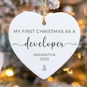 Peut inclure: Ornement de Noël blanc en forme de cœur avec une ficelle dorée. L'ornement porte l'inscription "MY FIRST CHRISTMAS AS A developer" en écriture noire, avec le nom "SAMANTHA" et l'année "2025" en dessous. Un petit sapin de Noël est en bas.