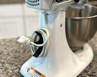 以下が含まれることがあります： 白いスタンドミキサーと銀色のボウル。ミキサーには黒いノブと、ベースに巻き付けられたコードがあります。ベースの警告ラベルには、「警告：器具が適切にアースされていない場合、深刻な感電の危険があります。アースされていないと、死亡または重傷を負う可能性があります。」と記載されています。
