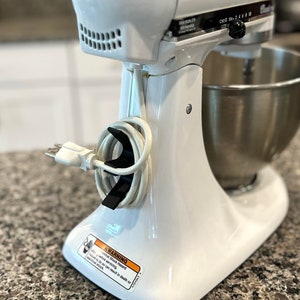 May include: A white stand mixer with a silver bowl. The mixer has a black knob and a cord wrapped around the base. The warning label on the base reads "WARNING:  A serious shock hazard exists if the appliance is grounded improperly.  Failure to ground can result in death or serious injury."