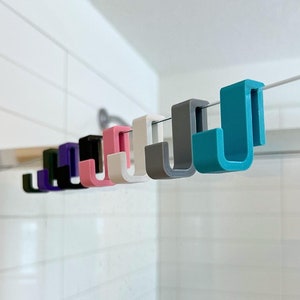 May include: A row of colorful, J-shaped hooks in various colors, including teal, gray, pink, and black, are suspended from a thin, silver wire. The hooks are evenly spaced and appear to be made of a solid material. The background is a white tiled wall.