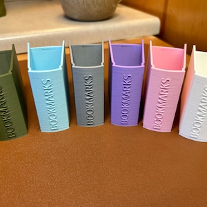 May include: Seven 3D printed bookmark holders in different colours: green, light blue, grey, purple, pink, and white. Each holder has the word "BOOKMARKS" embossed on the front.