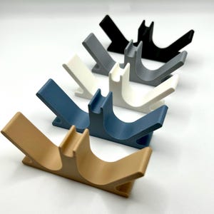 May include: Five 3D printed phone stands in different colours: beige, blue, white, grey, and black. The stands are shaped like a V with a curved section in the middle to hold a phone.