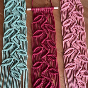 Puede incluir: Tres tapices de macramé en tonos turquesa, burdeos y rosa. Cada uno presenta un diseño de hojas y cuelga de una varilla de madera blanca. Los intrincados nudos crean un efecto texturizado, ideal para añadir un toque bohemio a cualquier espacio.