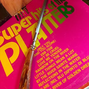 May include: A vintage record sharpener with a brown marbled handle and a silver metal shaft. The sharpener rests on a pink vinyl record album cover with the text "Super Hits of the Platters" in yellow and green.