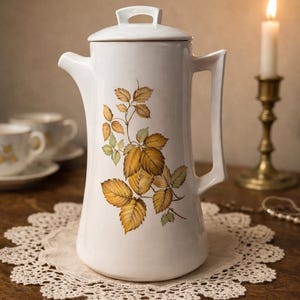 Peut inclure: Pichet à café en céramique blanche avec couvercle, anse et bec verseur. Décoré d'un motif botanique de feuilles jaunes et vertes. Posé sur une nappe en dentelle sur une table en bois, avec une bougie allumée en arrière-plan.