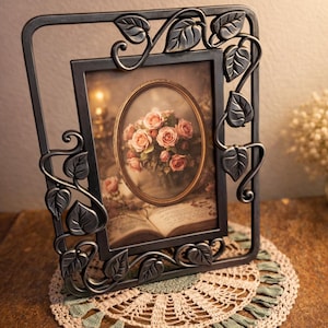 Puede incluir: Marco de fotos decorativo de metal gris oscuro con diseño floral. Contiene una imagen ovalada de rosas rosas y se apoya sobre un tapete de ganchillo. El marco tiene detalles de hojas y vides.