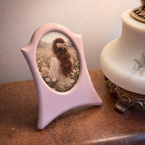 Peut inclure: Un cadre photo rose clair de forme ovale avec un design décoratif. Le cadre contient une photographie d'une personne dans un champ de fleurs. Une lampe vintage est en arrière-plan.