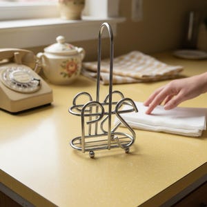 Może przedstawiać: Chromowany uchwyt na serwetki o dekoracyjnym wzorze, trzymający stos białych serwetek. Uchwyt stoi na żółtym blacie, z zabytkowym telefonem i cukiernicą we wzory kwiatowe w tle.