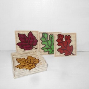 May include: A set of four square coasters and a matching holder, all featuring a leaf design. The coasters and holder are made of a beige material with cross-stitch leaves in shades of red, green, and gold.