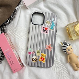 Pode incluir: Uma capa de telefone prateada e preta decorada com adesivos, incluindo a Estátua da Liberdade e um ursinho de pelúcia. A imagem também mostra um bálsamo labial rosa, um frasco de perfume e uma pequena estatueta.