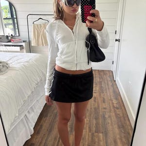 May include: A white zip-up hoodie with a drawstring hood, a black mini skirt with white side stripes, and white flip-flops. The outfit is accessorized with black sunglasses, gold hoop earrings, a black shoulder bag, and a black phone case.