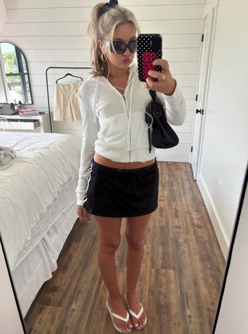 May include: A white zip-up hoodie with a drawstring hood, a black mini skirt with white side stripes, and white flip-flops. The outfit is accessorized with black sunglasses, gold hoop earrings, a black shoulder bag, and a black phone case.