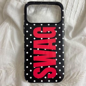 May include: A black smartphone case featuring a white polka dot pattern and the word SWAG printed in large, bold red capital letters vertically across the center. The case has a rectangular cutout at the top for a camera lens and a black border.