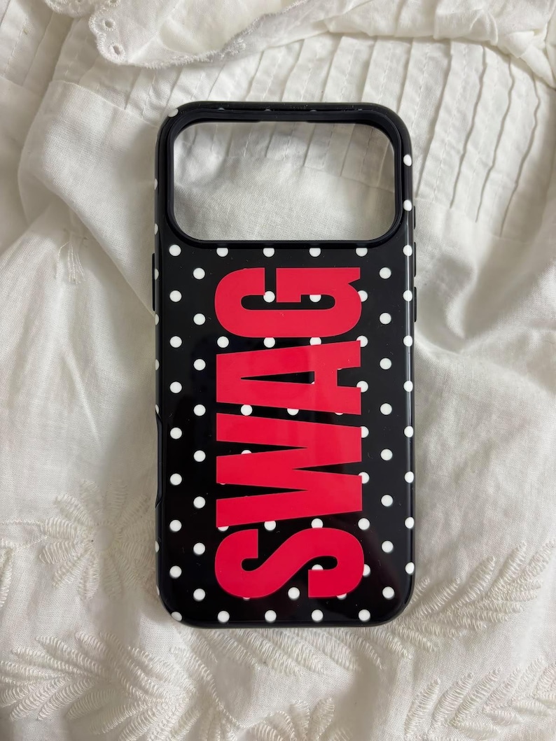 May include: A black smartphone case featuring a white polka dot pattern and the word SWAG printed in large, bold red capital letters vertically across the center. The case has a rectangular cutout at the top for a camera lens and a black border.