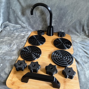 May include: A black faucet with a curved spout is mounted on a bamboo cutting board. The board also holds black circular trivets, star-shaped knobs, and a handle. The items are arranged on a gray, textured surface.