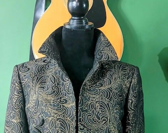 May include: A black jacket featuring a prominent gold-colored paisley pattern throughout the fabric. The garment has a structured high collar, long sleeves, and an open front design, displayed on a black mannequin torso against a green background.
