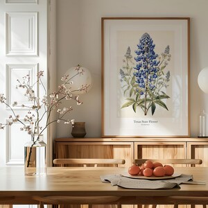 May include: A framed botanical print of the Texas State Flower, a bluebonnet, hangs above a wooden sideboard. A vase of flowering branches and a bowl of oranges sit on a wooden dining table. The room is decorated in neutral tones.