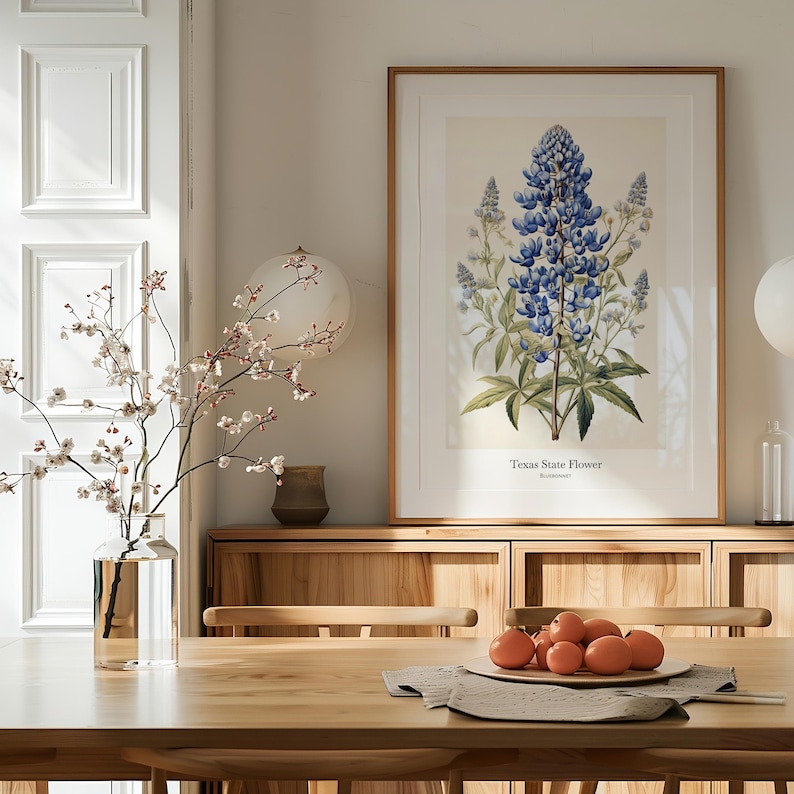 May include: A framed botanical print of the Texas State Flower, a bluebonnet, hangs above a wooden sideboard. A vase of flowering branches and a bowl of oranges sit on a wooden dining table. The room is decorated in neutral tones.