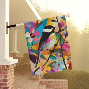 Pode incluir: Uma bandeira de jardim colorida com um pássaro estilizado em preto e branco empoleirado em um galho com flores e folhas coloridas. A bandeira é feita de tecido e tem um design vibrante e abstrato.