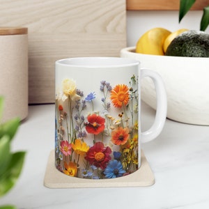 May include: White ceramic mug with a colourful floral design, including yellow, orange, red, and blue flowers. The mug rests on a beige coaster. A white handle is on the right. The mug's design is reminiscent of a wildflower meadow.