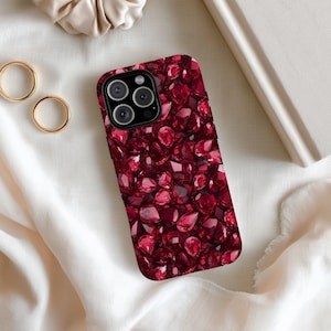 May include: A smartphone case with a vibrant pattern of deep red gemstones. The case has a black border and a camera cutout. The gemstones are various shapes and sizes, creating a textured appearance. Gold rings and a beige scrunchie are in the background.